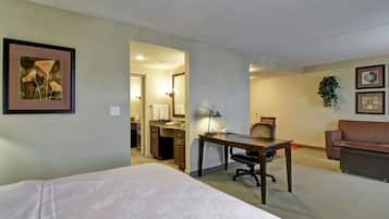 A hotel room with a bed, desk, chair, and a small seating area. There is a kitchenette in the background.