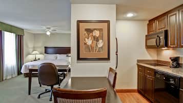 A hotel room with a bed, a dining table with chairs, a kitchen area with a microwave and dishwasher, and a ceiling fan.