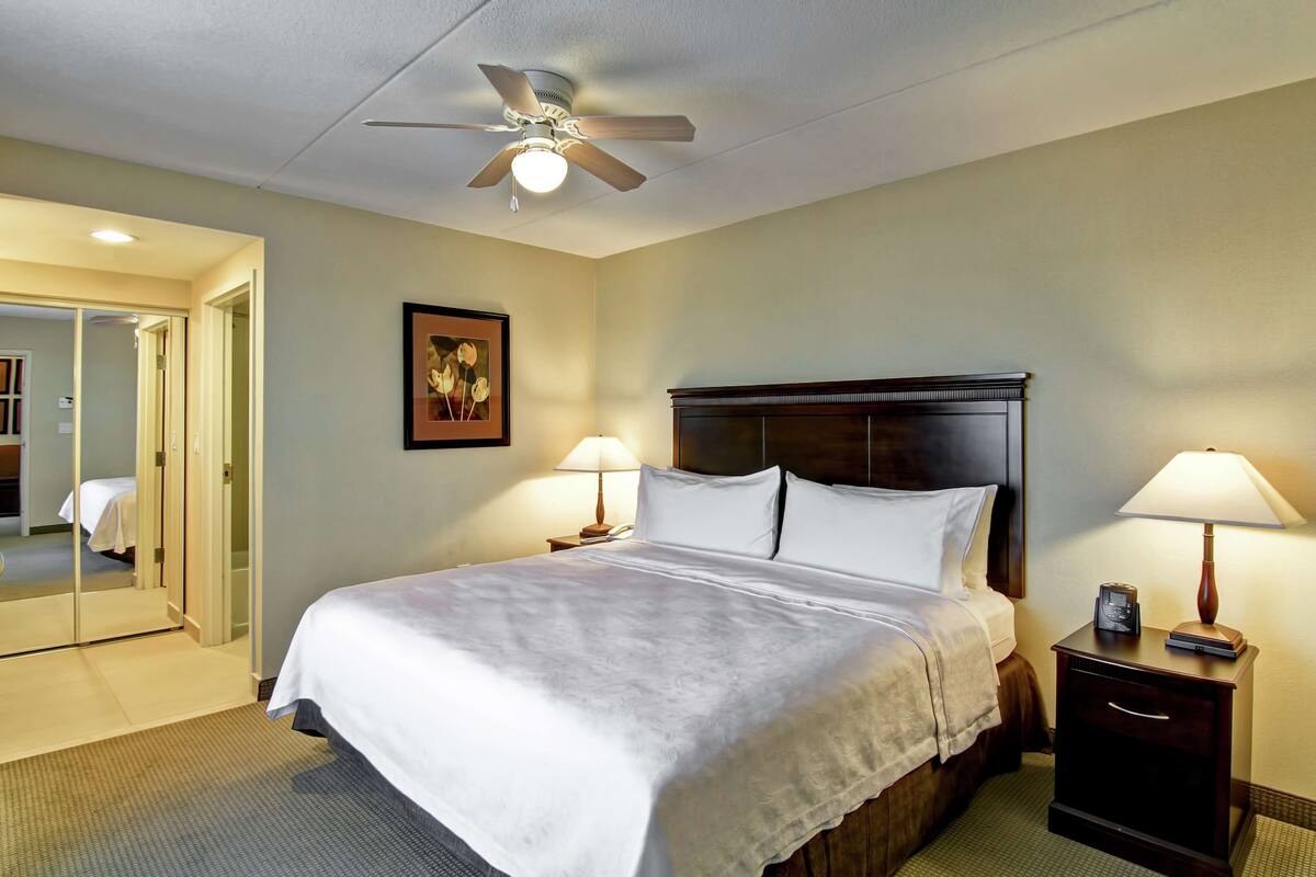 Une chambre d’hôtel avec un lit, des lampes de chevet, un ventilateur de plafond, un tableau encadré au mur et un miroir.