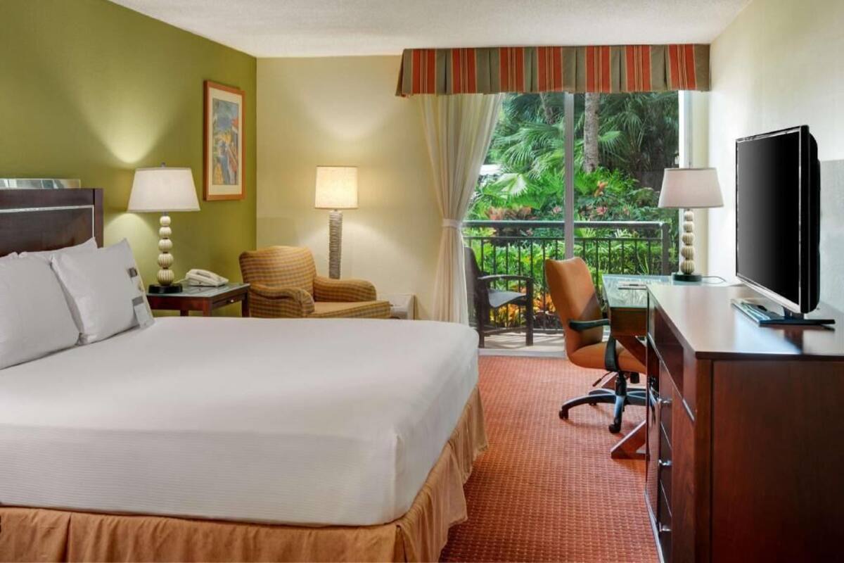 Une chambre d’hôtel équipée d’un lit, d’un bureau, d’une chaise, d’une télévision et d’un balcon offrant une vue sur la verdure.