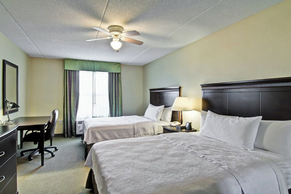 Une chambre d’hôtel avec deux lits, un bureau, une chaise, une lampe et un ventilateur de plafond.