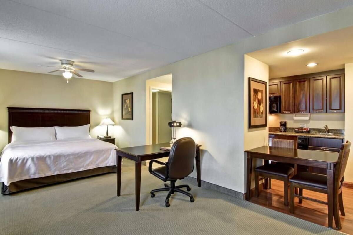 Une chambre d’hôtel équipée d’un lit, d’un bureau, d’une chaise, d’une kitchenette et d’un ventilateur de plafond.