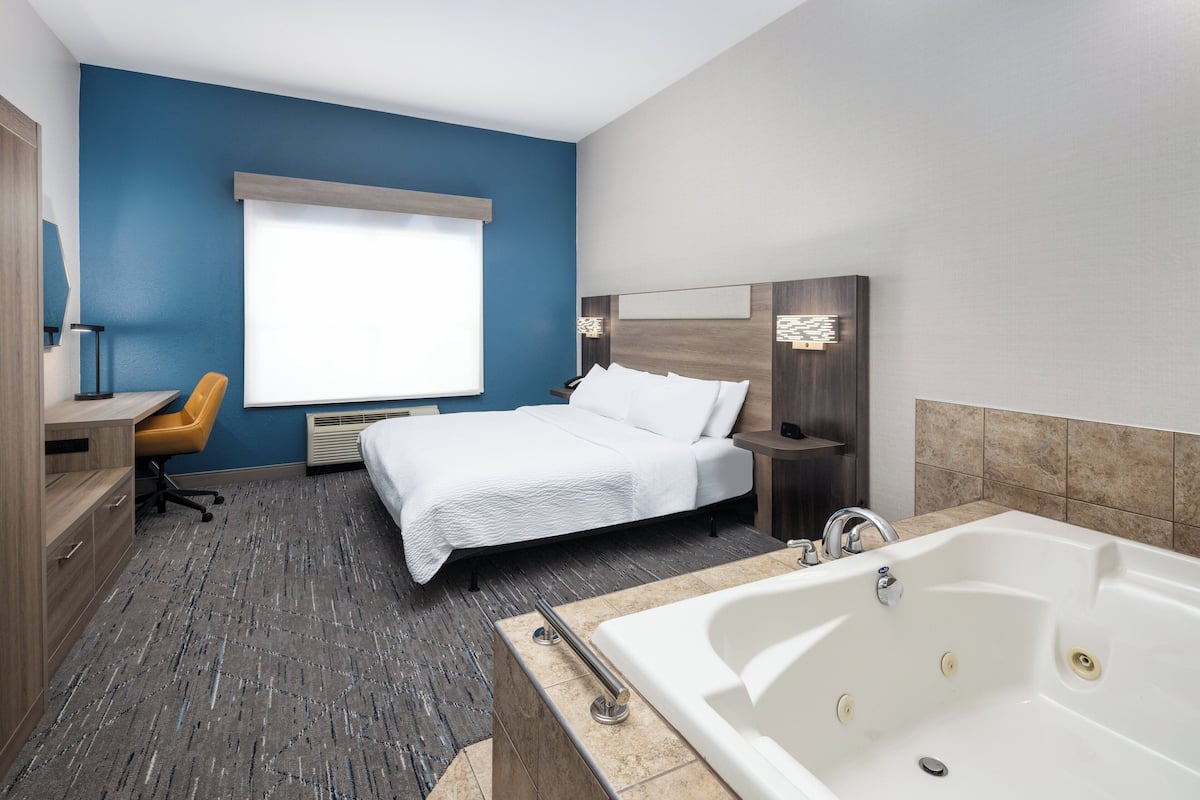 A modern hotel room with a large bed, a desk, a chair, a bathtub, and a window with blinds.