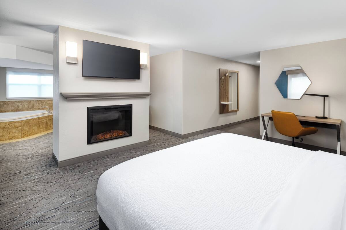 A hotel room with a bed, a TV mounted on the wall, a fireplace, a desk with a chair, and a large mirror.