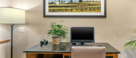A hotel room with a desk, computer, chair, lamp, and a framed picture on the wall.