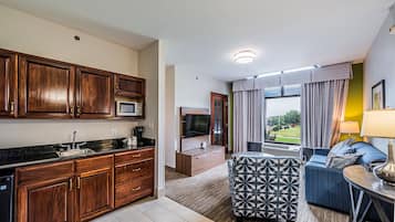 A modern kitchen with wooden cabinets, a microwave, and a sink. There is a living area with a sofa and a patterned ottoman. A television is mounted on the wall, and a window with curtains is visible.