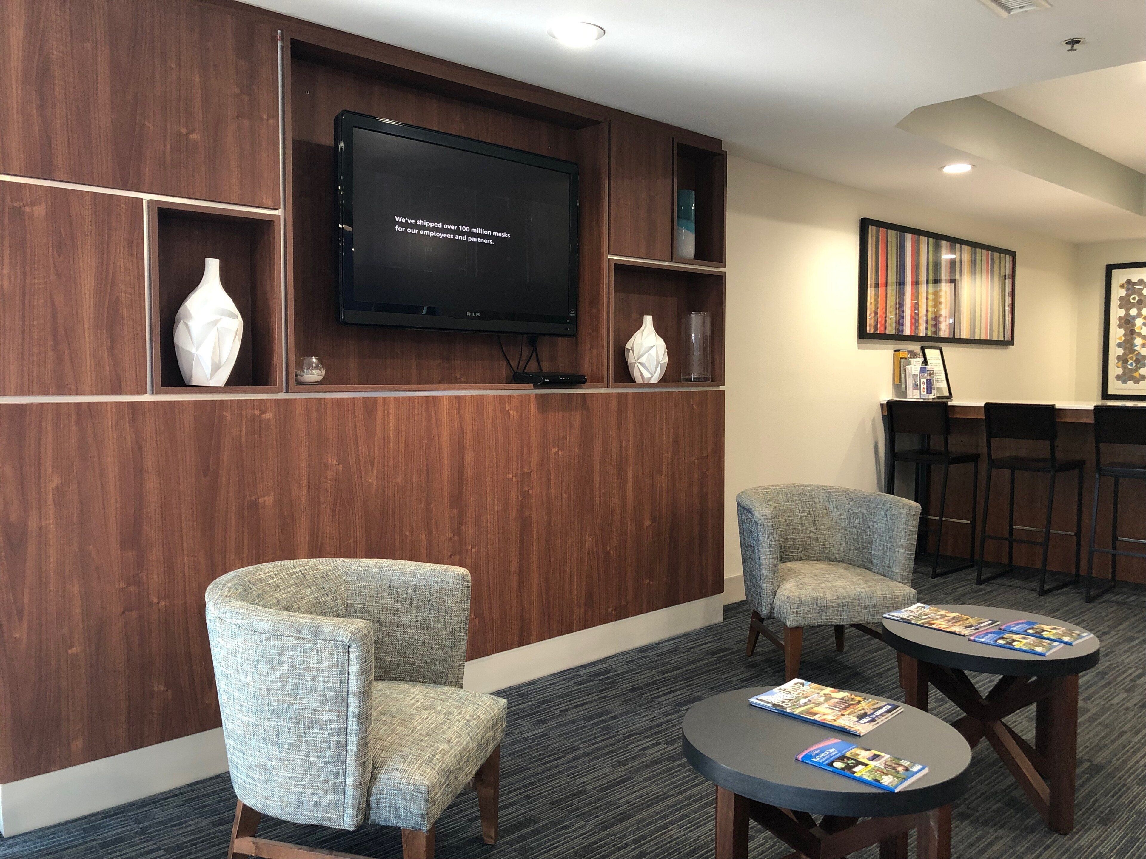A lobby with two upholstered chairs, a round table with magazines, a flat-screen TV on a wooden wall, and a bookshelf with decorative items.