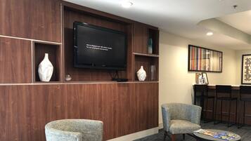 A lobby with two upholstered chairs, a round table with magazines, a flat-screen TV on a wooden wall, and a bookshelf with decorative items.