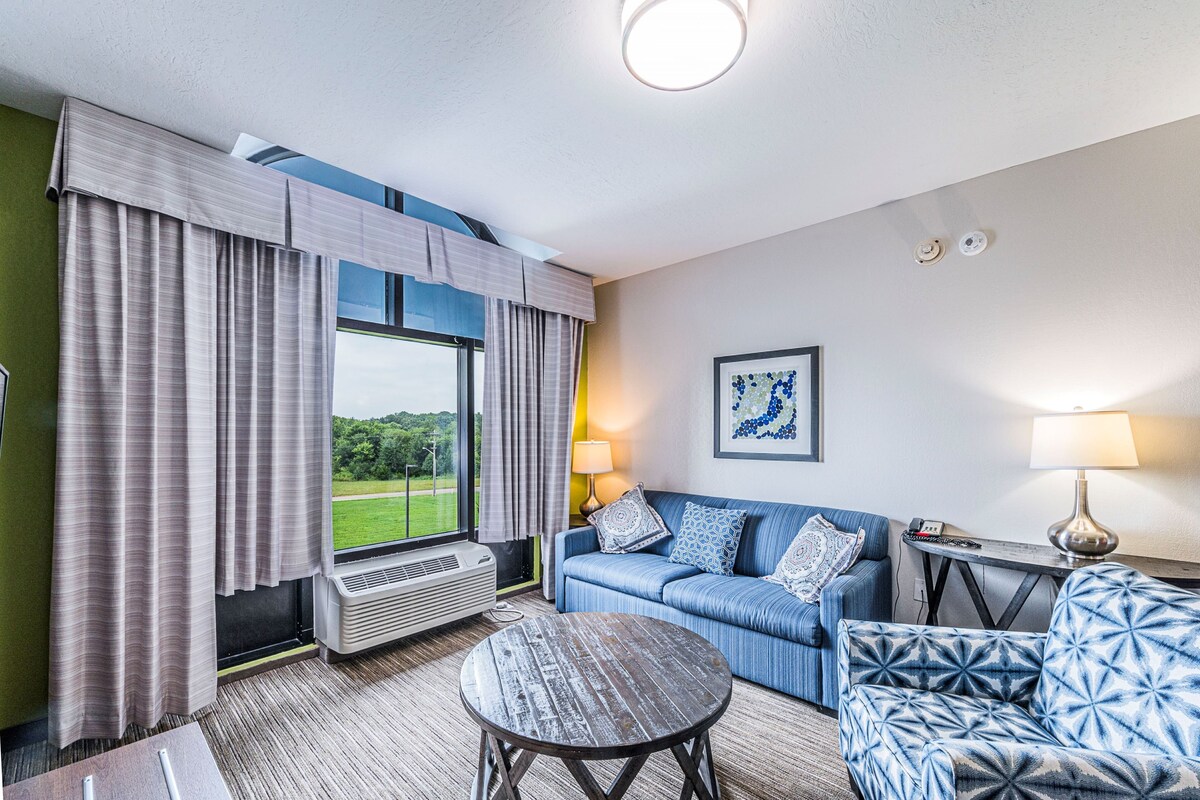 A hotel room with a large window, a sofa, a patterned armchair, a small round table, and a lamp.