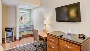 A hotel room with a desk, chair, TV, and a small kitchenette with an oven.