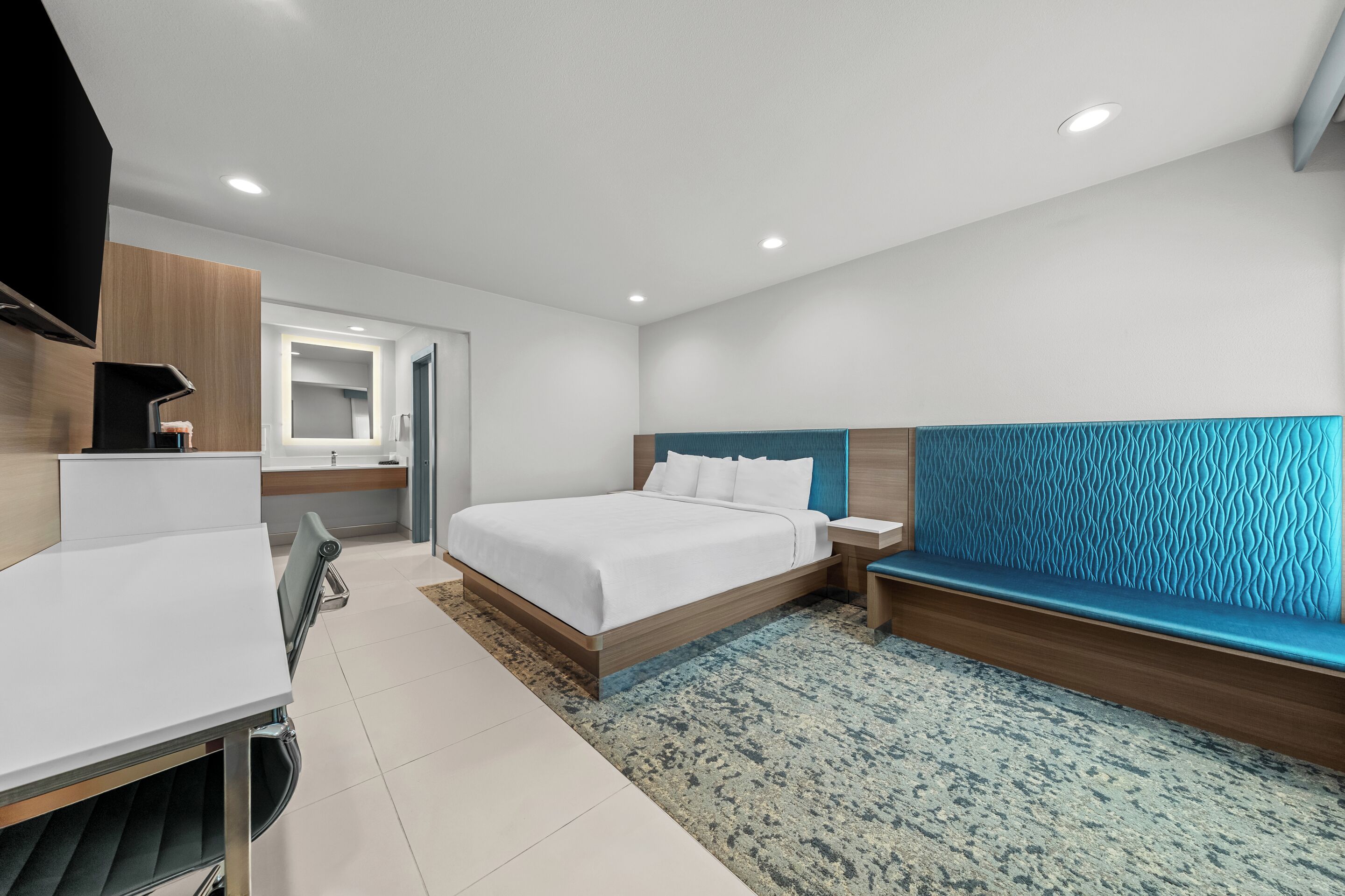 A modern hotel room with a bed, bedside table, and a seating area with a blue upholstered headboard.