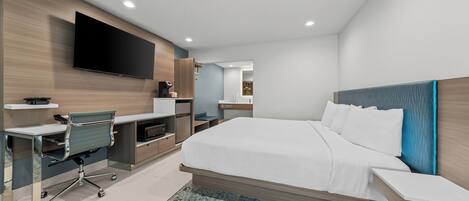 A modern hotel room with a large bed, a desk with a chair, a flat-screen TV mounted on the wall, and a small kitchenette area.