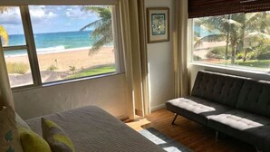 A bedroom with a large window offering a beach view, a bed with pillows, and a grey sofa.