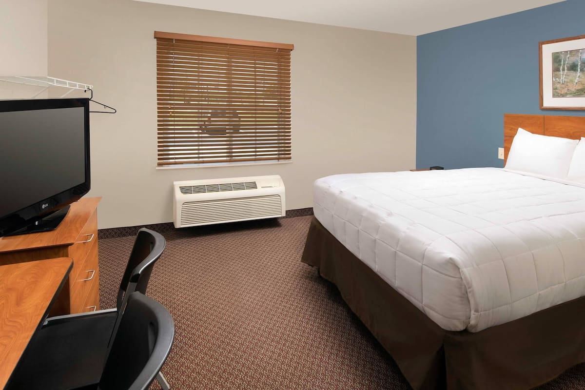 A hotel room with a bed, a desk with a television, a chair, a window with blinds, and a wall-mounted air conditioner.