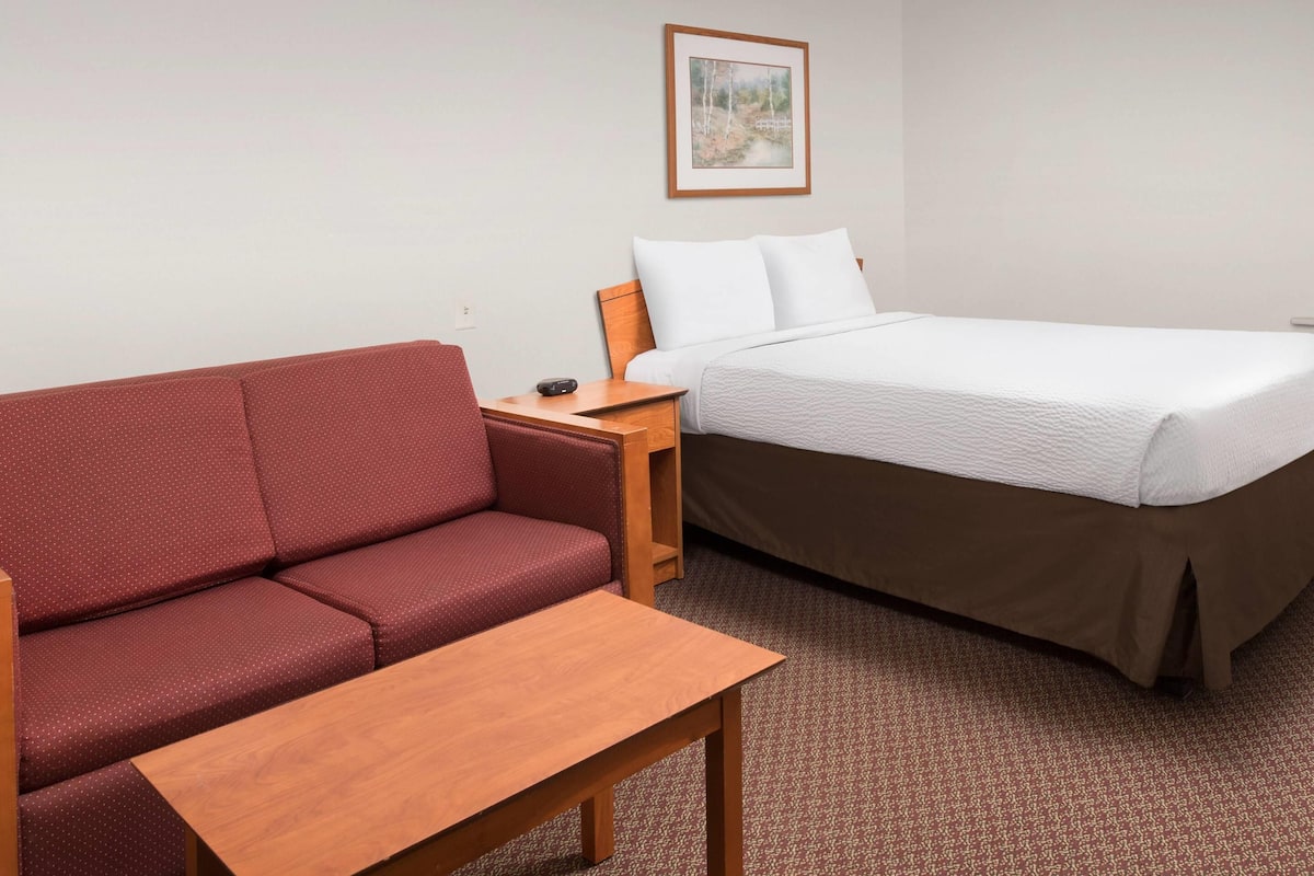 A hotel room with a red sofa, a wooden table, a bed with white bedding, and a small nightstand with a lamp.