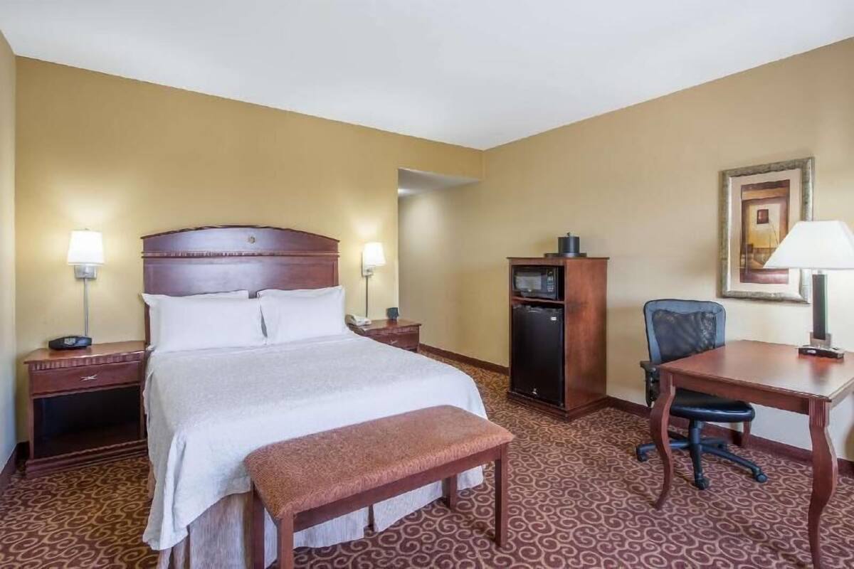 A hotel room with a bed, bedside tables, a desk, a chair, a refrigerator, and a lamp.