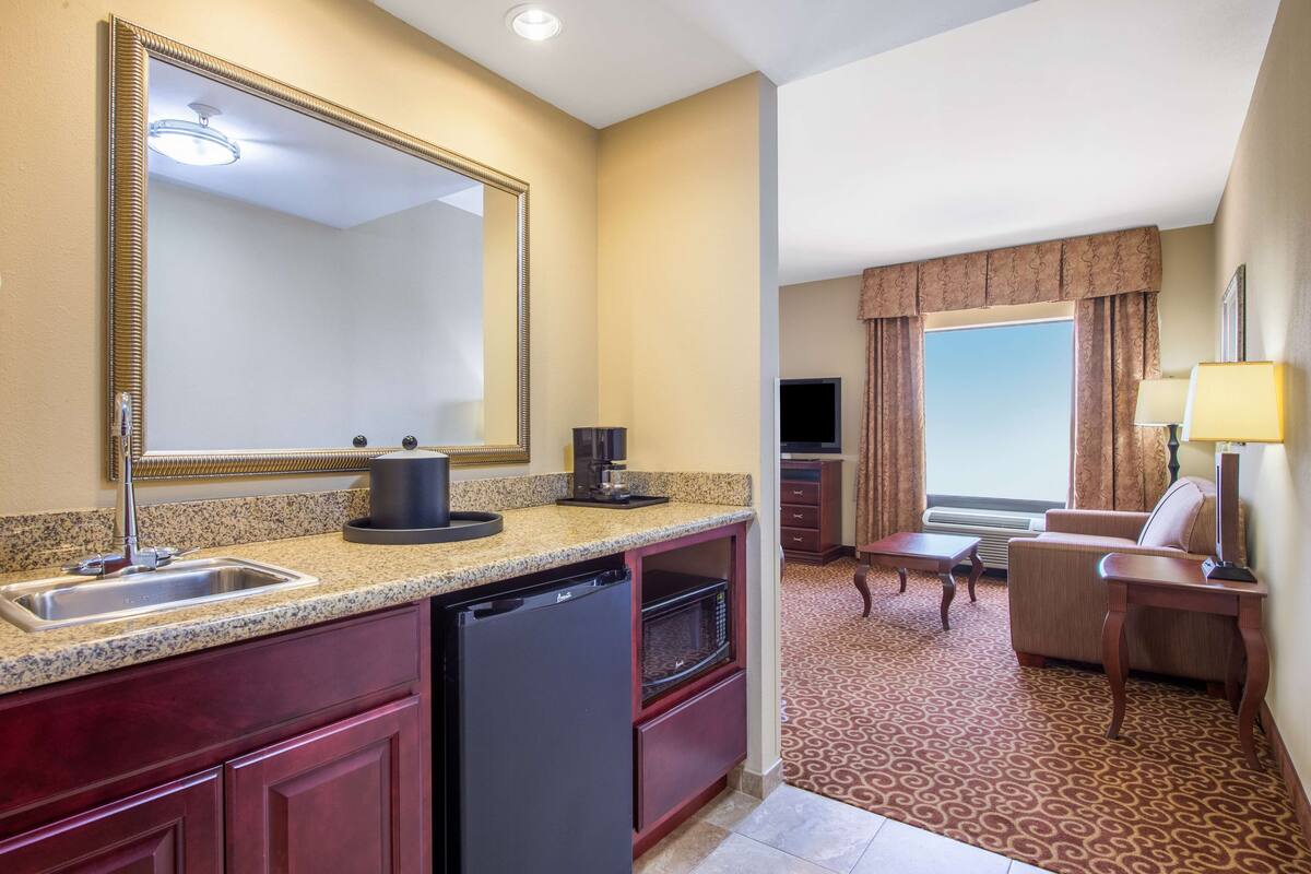 A hotel room with a kitchenette, a large mirror, a flat-screen TV, a sofa, and a patterned carpet.