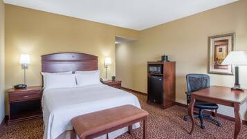 Quarto de hotel com uma cama grande, mesas de cabeceira com abajures, uma escrivaninha com cadeira, um frigorífico e um tapete estampado.
