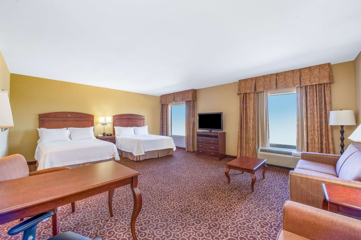 A hotel room with two beds, a TV, a desk, a chair, a sofa, and a window with curtains.