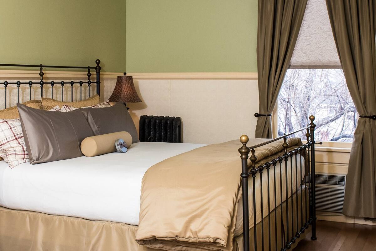 A bedroom with a bed, a radiator, a window with curtains, and a lamp.