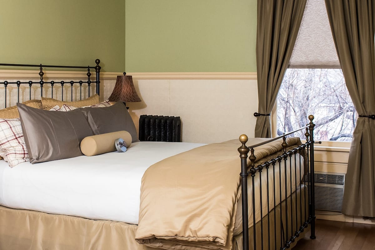 A bedroom with a bed, a radiator, a window with curtains, and a lamp.