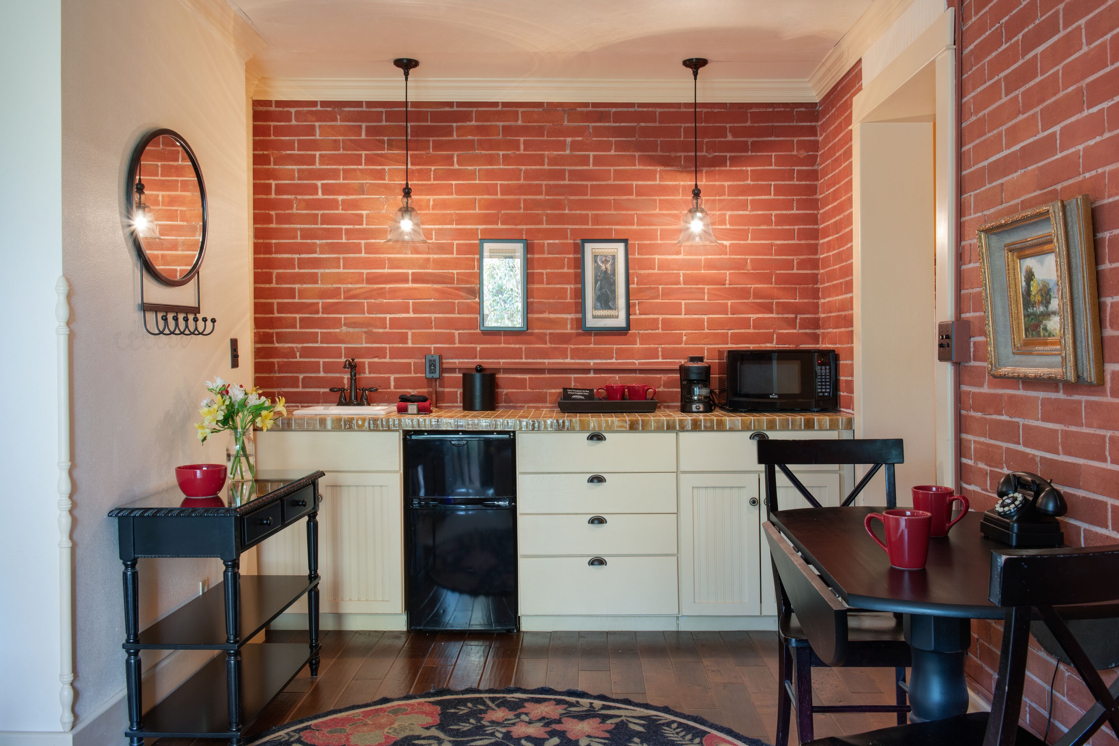 Una cocina compacta con pared de ladrillo, muebles blancos, un refrigerador negro y una zona de comedor con una mesa y sillas.