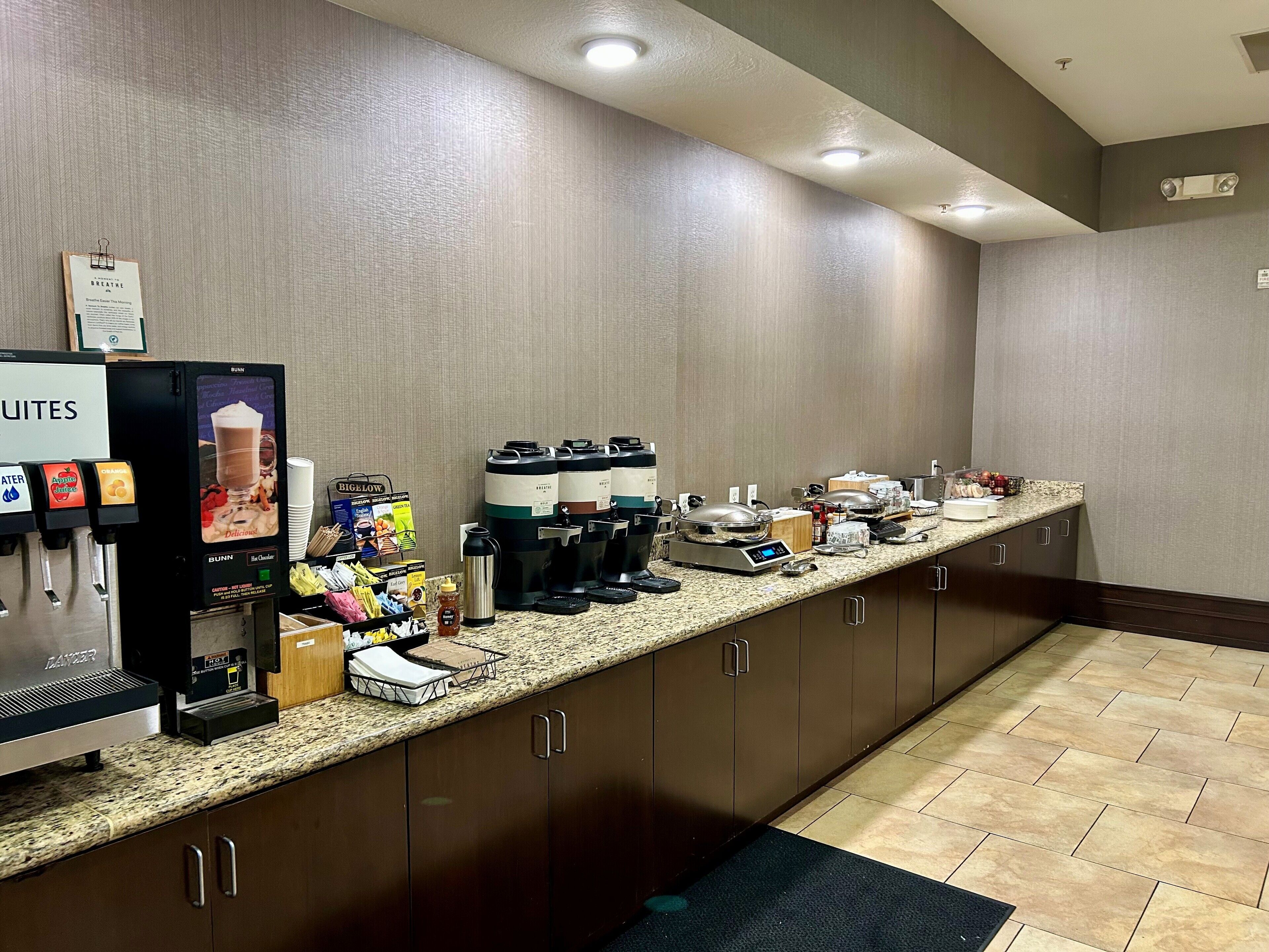 Una zona de cocina de hotel con estación de café, dispensadores de bebidas y diversos alimentos sobre la encimera.