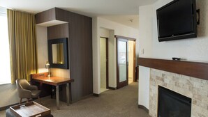 A hotel room with a flat-screen TV, a desk with a chair, a fireplace, and a large window with curtains.