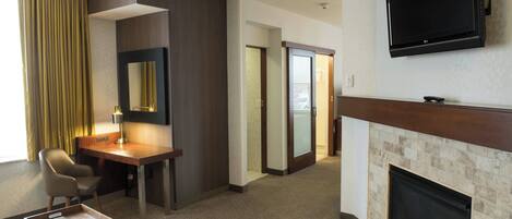 A hotel room with a flat-screen TV, a desk with a chair, a fireplace, and a large window with curtains.