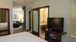 A hotel room with a bed, a television on a stand, a bathroom with a mirror, and a sofa in the corner.