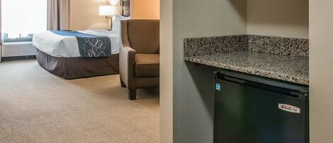 A hotel room with a bed, a chair, a desk, and a refrigerator.
