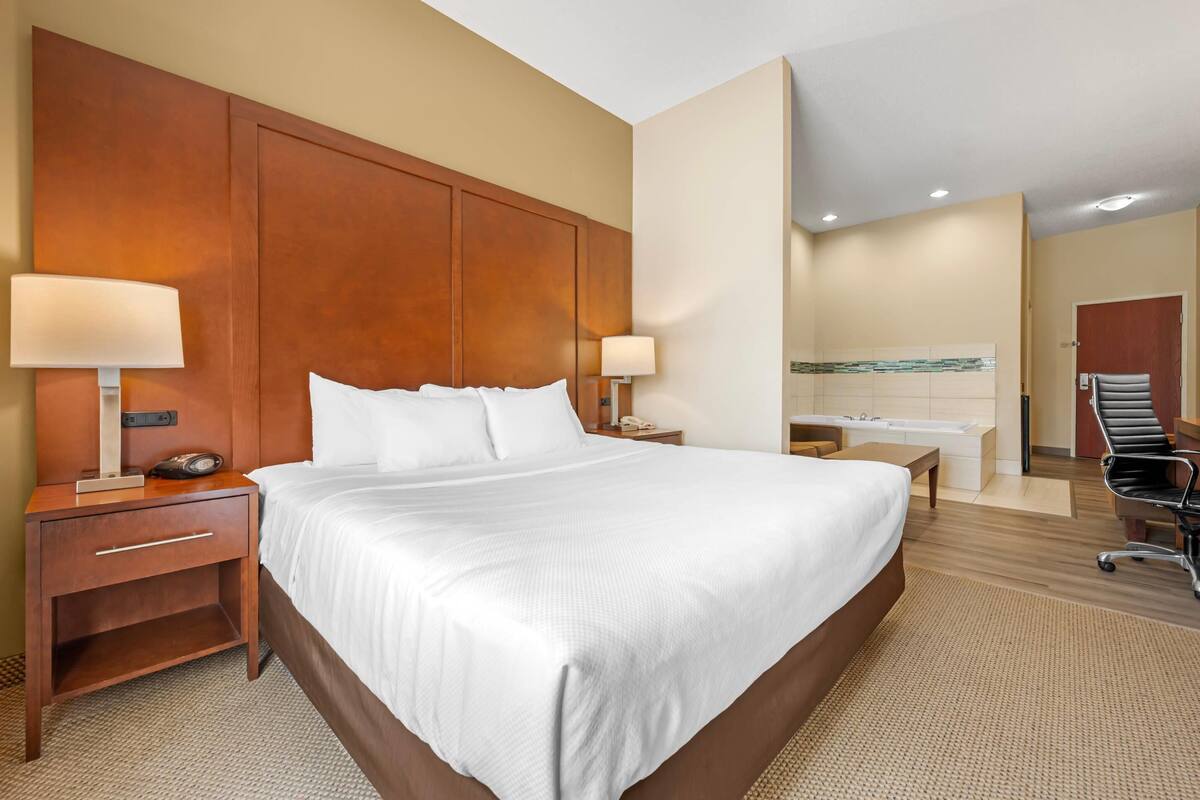 A hotel room with a large bed, two bedside tables with lamps, a desk with a chair, and a bathroom visible through an open door.