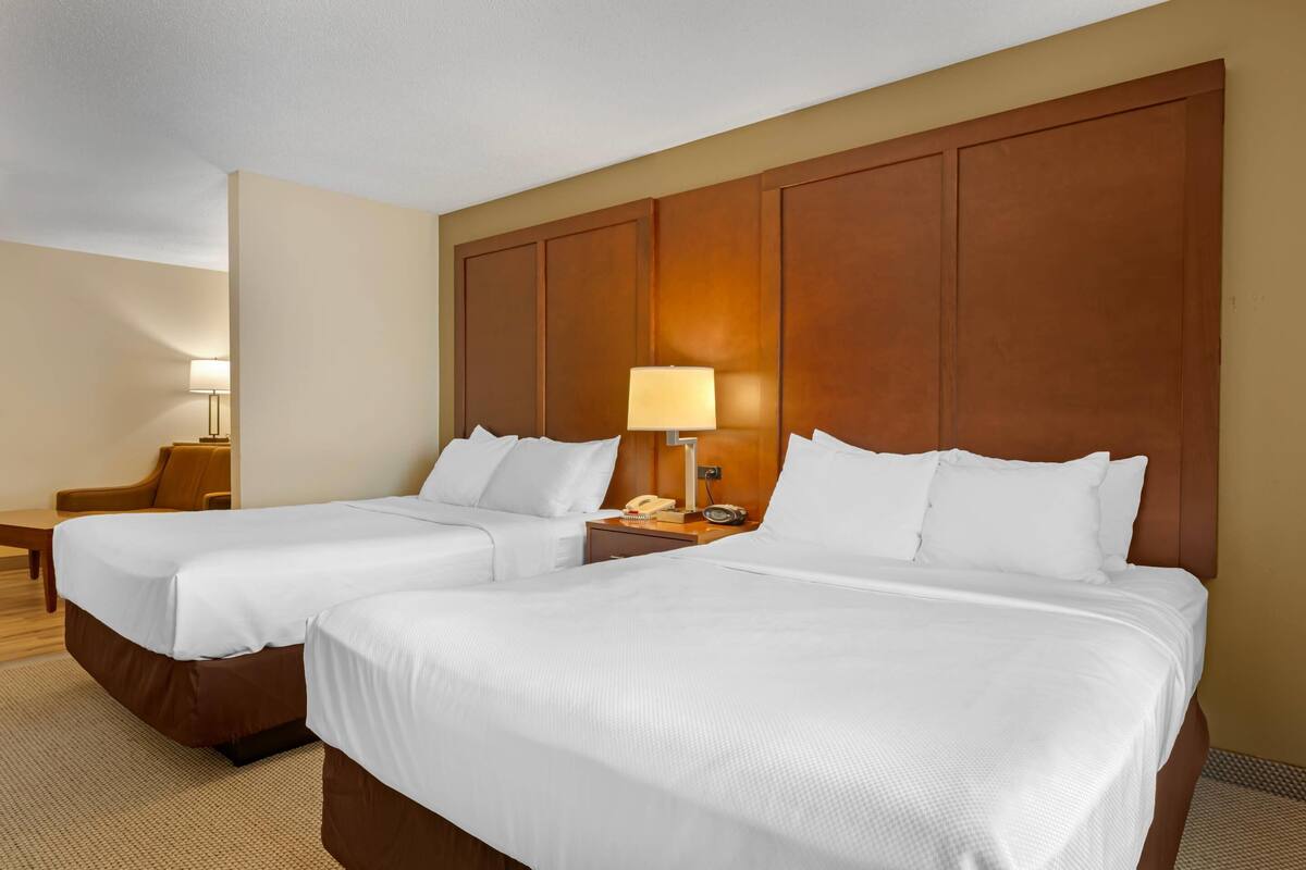 A hotel room with two beds, a wooden headboard, a bedside table with a lamp, and a chair.