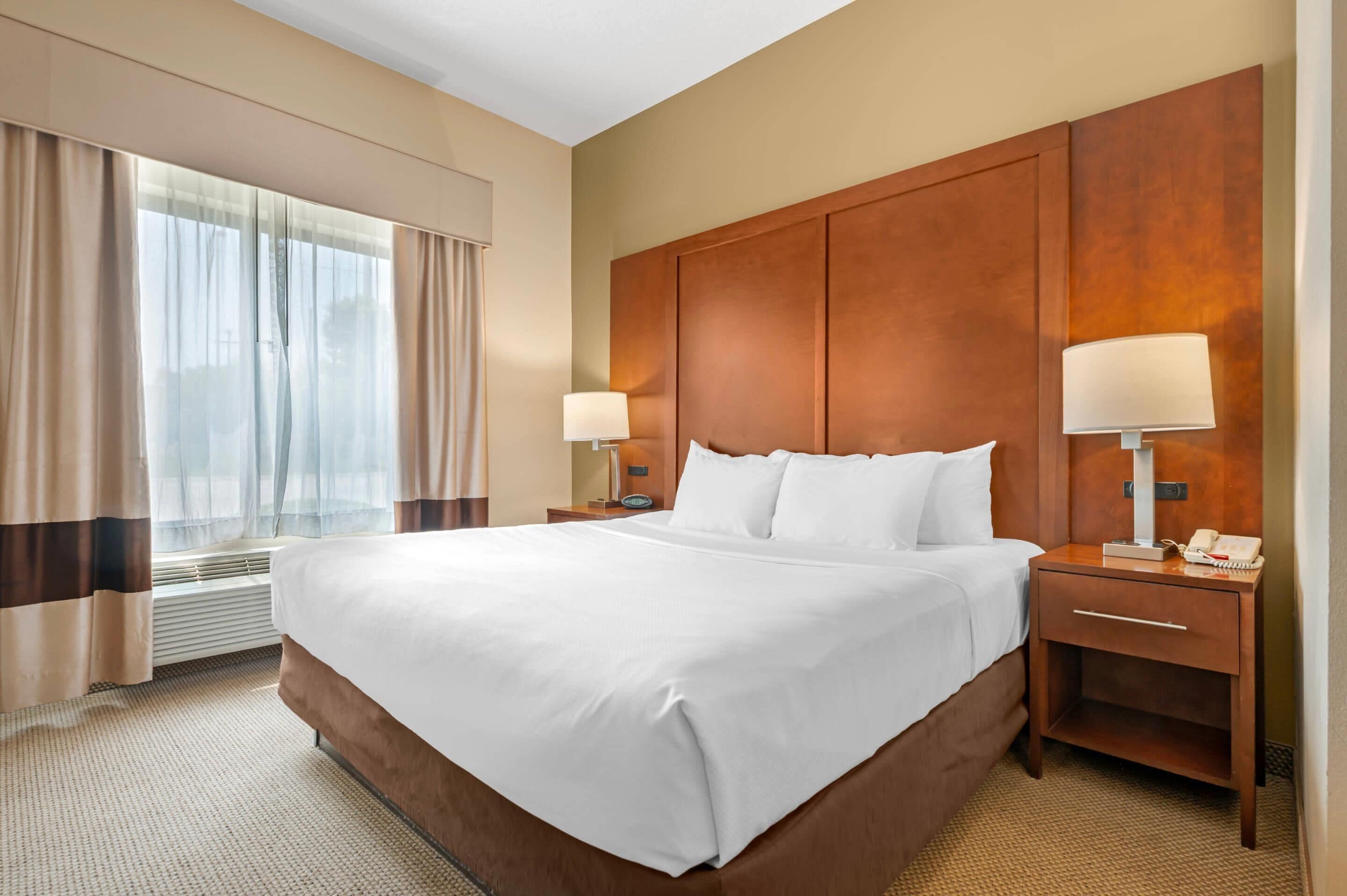 A hotel room with a large bed, two bedside tables with lamps, a window with curtains, and a wooden headboard.