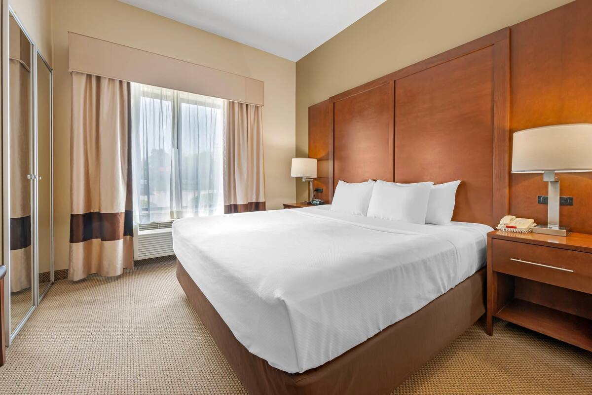 A hotel room with a large bed, a wooden headboard, a bedside table with a lamp, and a window with curtains.