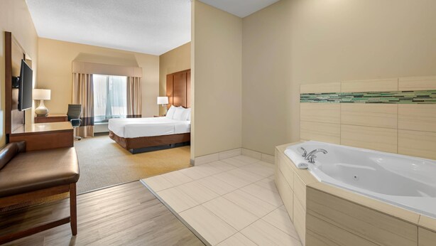 Habitación de hotel con una cama grande, una zona de estar independiente con una silla, un escritorio, un televisor, un baño con bañera y un ventanal con cortinas.