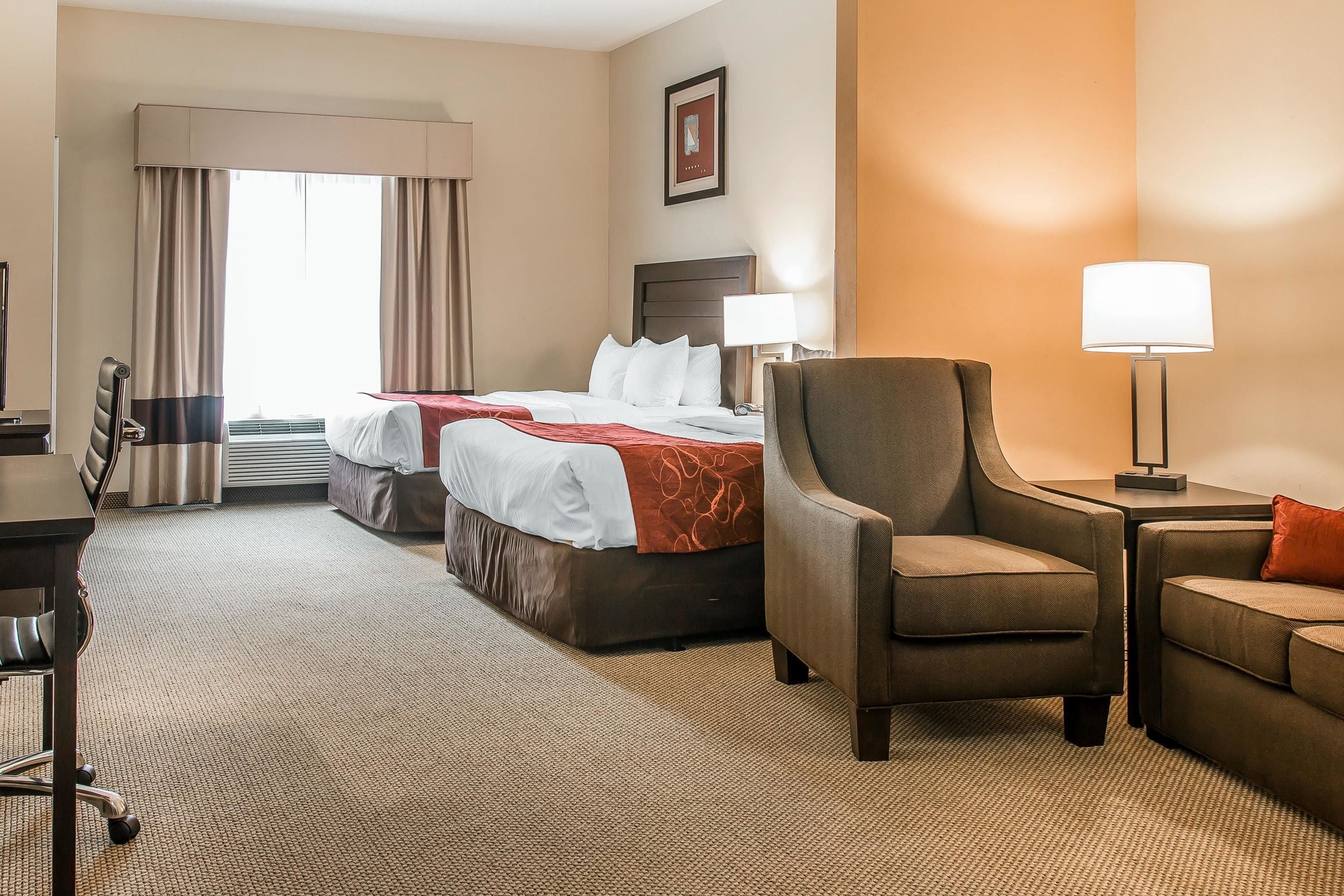 A hotel room with two beds, a desk, a chair, a sofa, and a lamp.