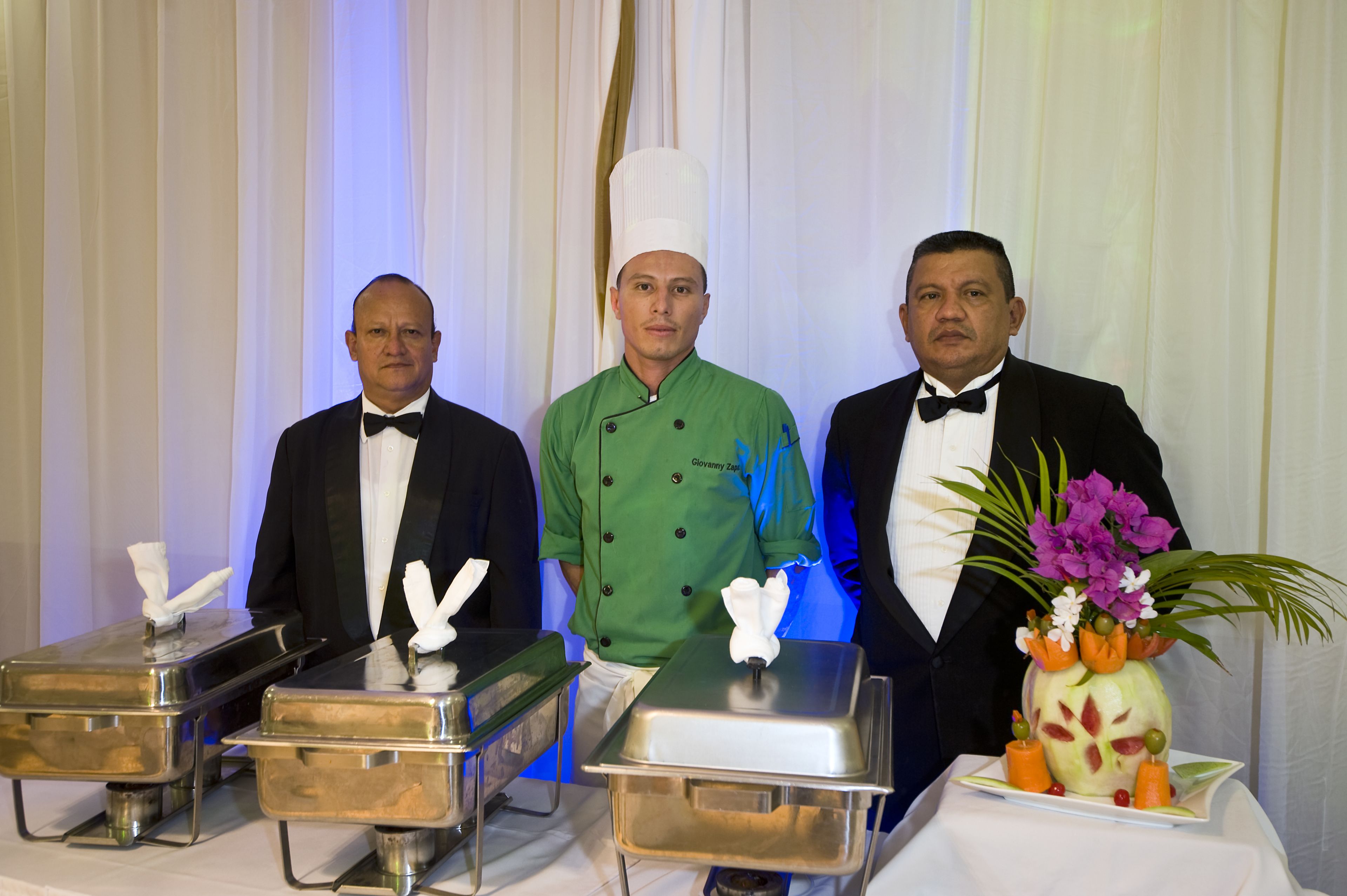 Three waitstaff in formal attire standing behind buffet stations with covered food containers.