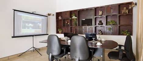 A conference room with a projector screen, a desk with a laptop, and a wall-mounted shelf with decorative items.