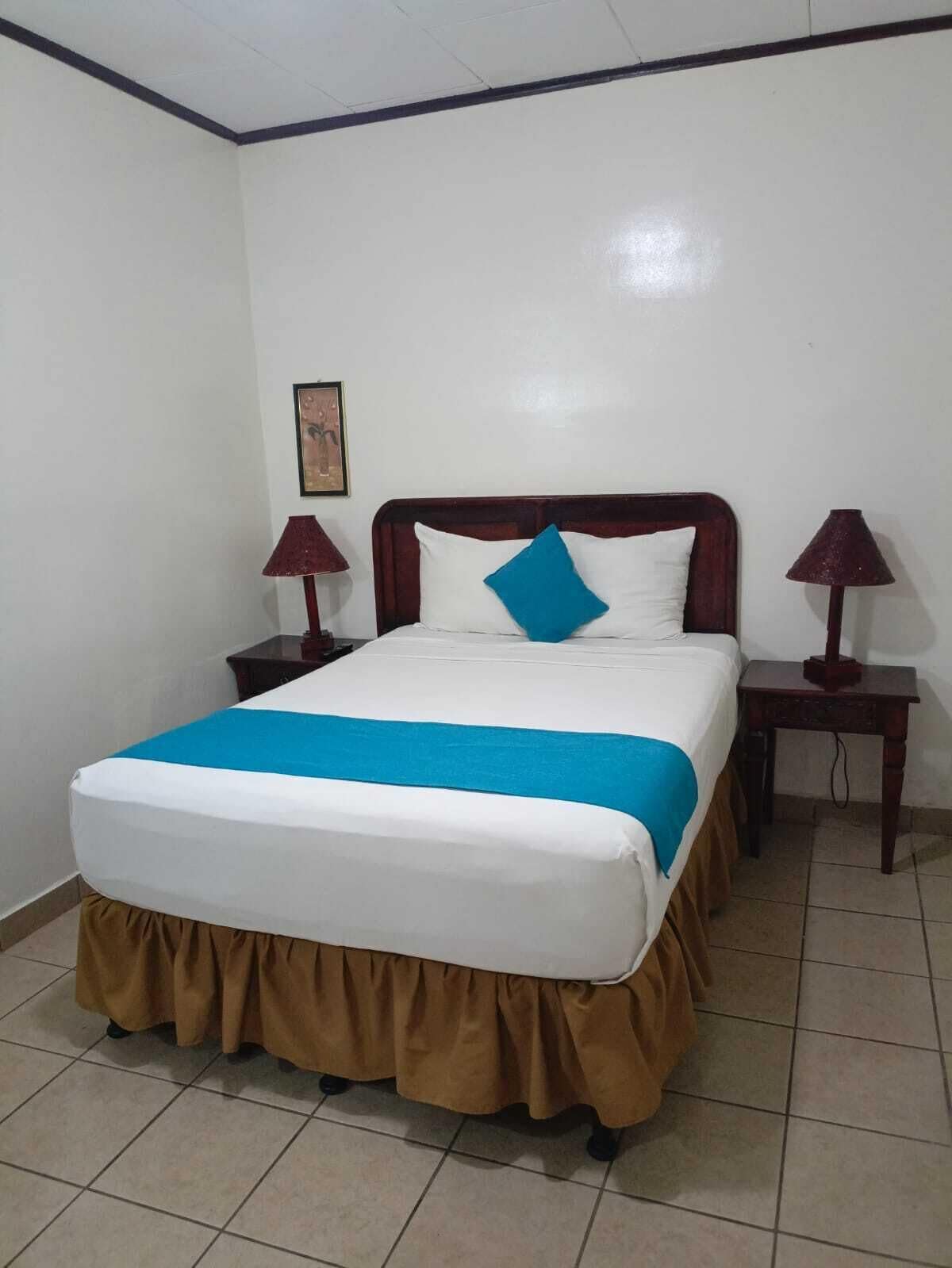 Una cama bien hecha con una colcha azul y falda marrón, dos mesitas de noche con lámparas, y un cuadro enmarcado en la pared.