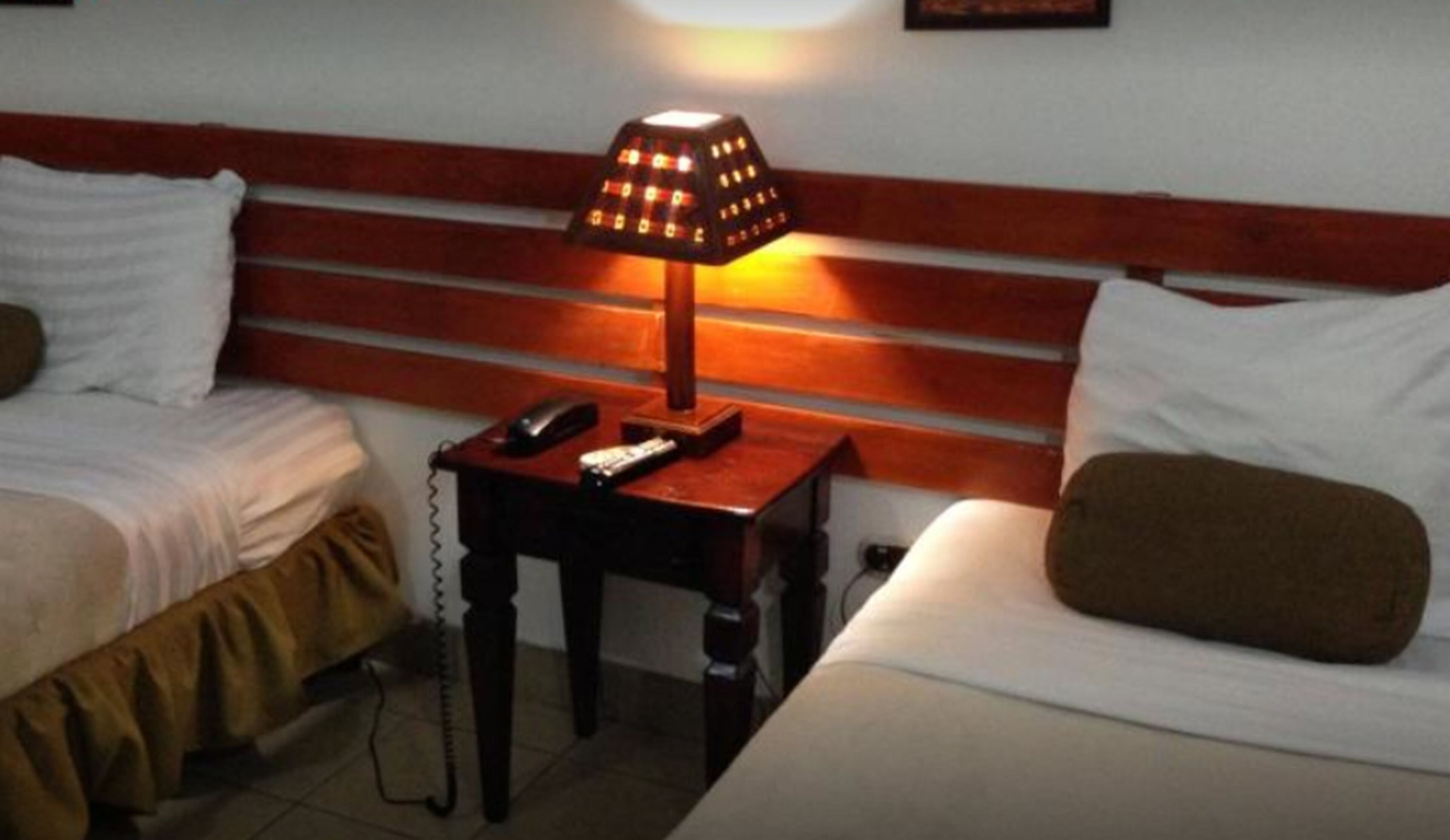 A hotel room with two single beds, a wooden headboard, a bedside table with a lamp, a telephone, and a remote control.