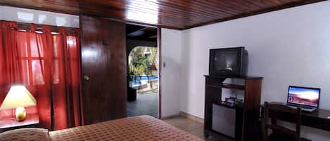 Habitación de hotel con una cama, un escritorio con computadora, televisión y puerta que da a un espacio exterior.