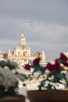 Um edifício histórico com várias cúpulas e uma cruz no topo, visto de uma varanda com vasos de plantas em primeiro plano.