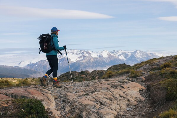 A hiker on a rocky trail with a backpack and trekking poles, surrounded by mountains and sparse vegetation.