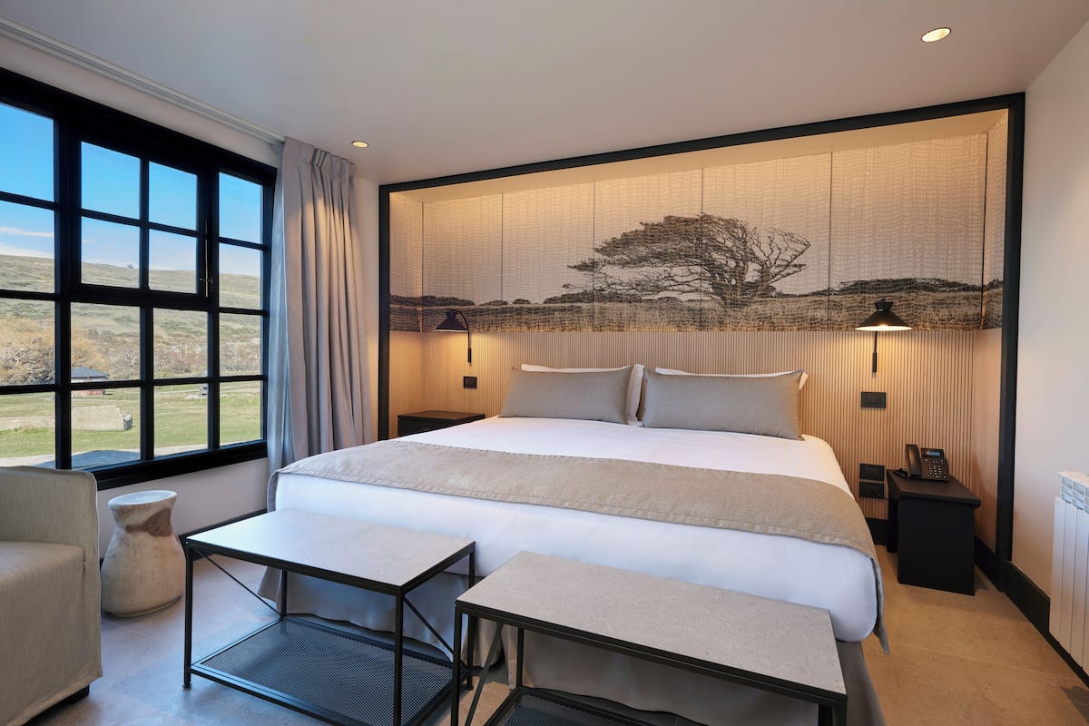 Ein modernes Hotelzimmer mit einem großen Bett, Nachttischen, einem Sessel und Blick ins Freie durch ein großes Fenster.