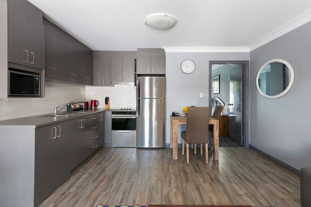 Een moderne keuken met grijze kastjes, roestvrijstalen apparatuur, een houten eettafel en een klok aan de muur.