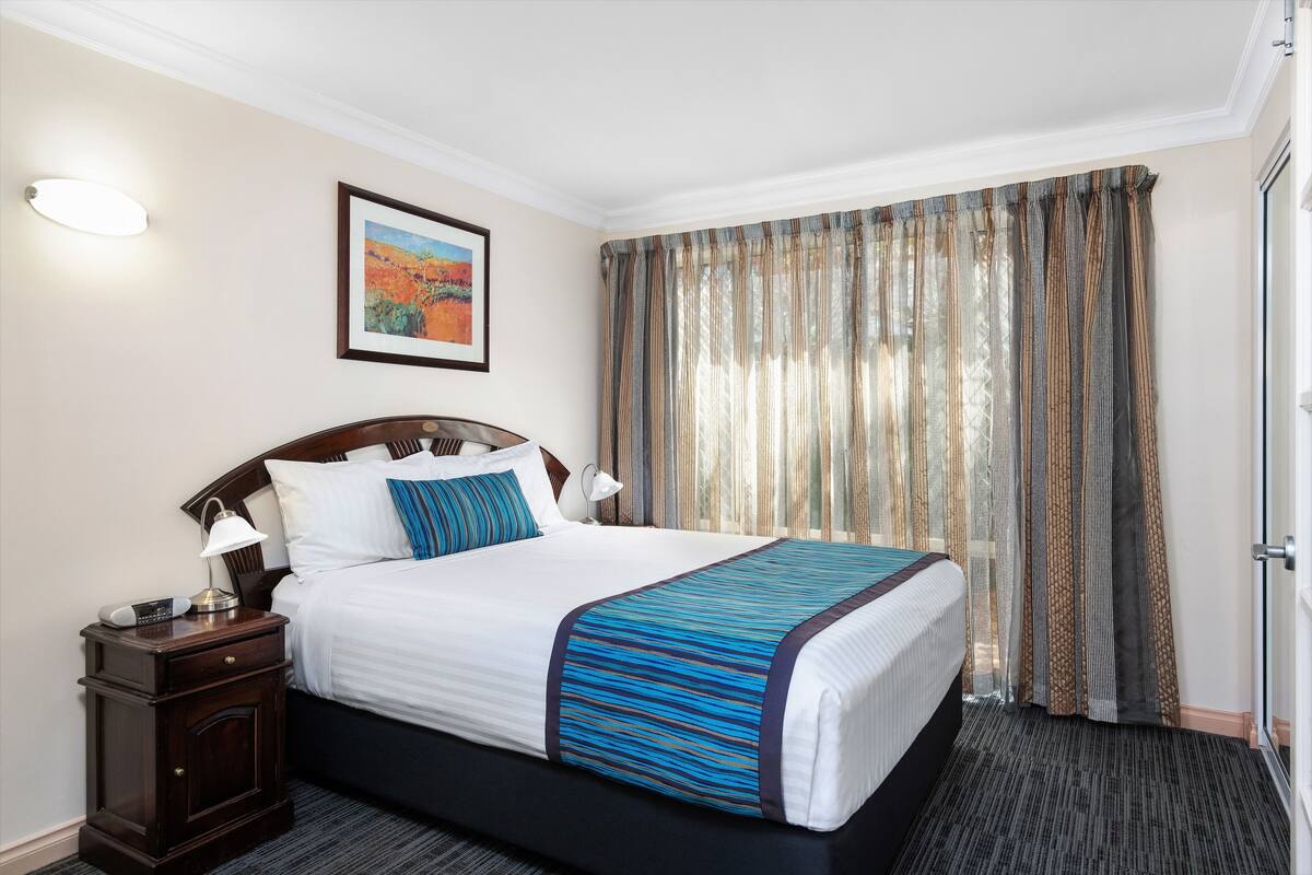 Een hotelkamer met een groot bed, een nachtkastje met een lamp, een ingelijst schilderij aan de muur en een raam met gordijnen.