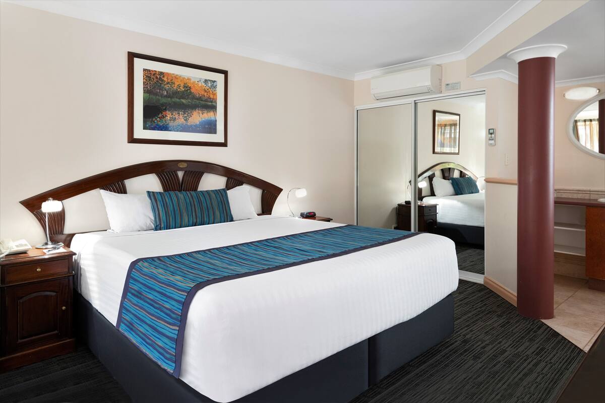 Een hotelkamer met een groot bed, nachtkastje, bureau, spiegel en een schilderij van een meer aan de muur.