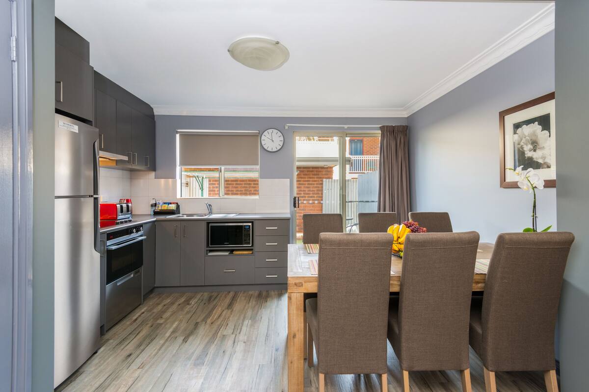 Een moderne keuken met grijze kastjes, een roestvrijstalen koelkast en een houten eettafel met vier stoelen.