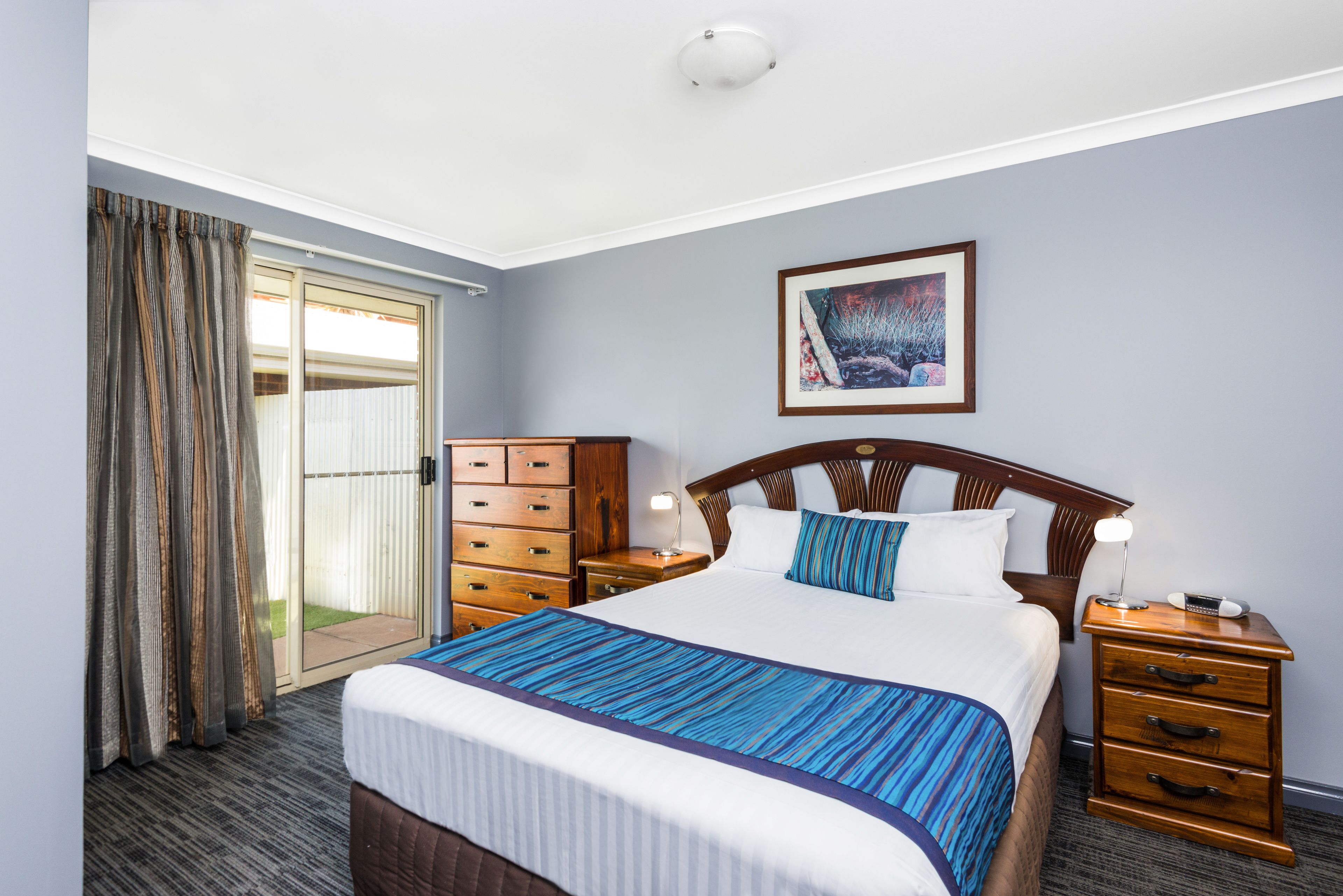 Une chambre d’hôtel avec un grand lit, des tables de chevet en bois, une commode, une porte-fenêtre coulissante et un tableau encadré au mur.